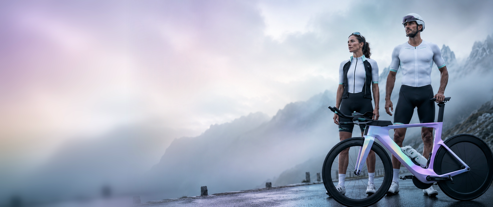 Two cyclists stand beside an iridescent bike on a misty mountain road.